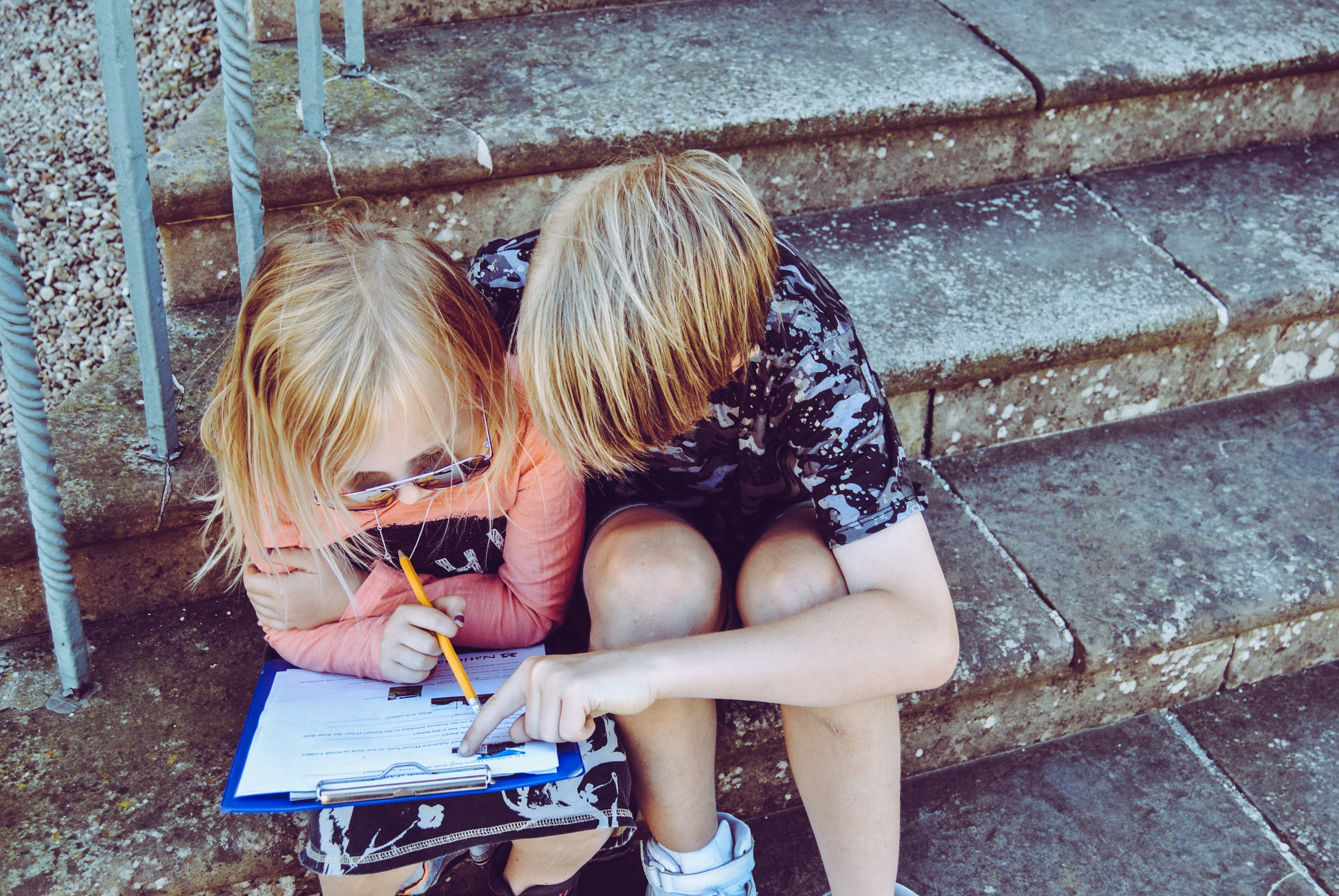 A girl and a boy sitting on a step, the girl holding a clipboard and the boy pointing to something on the clipboard