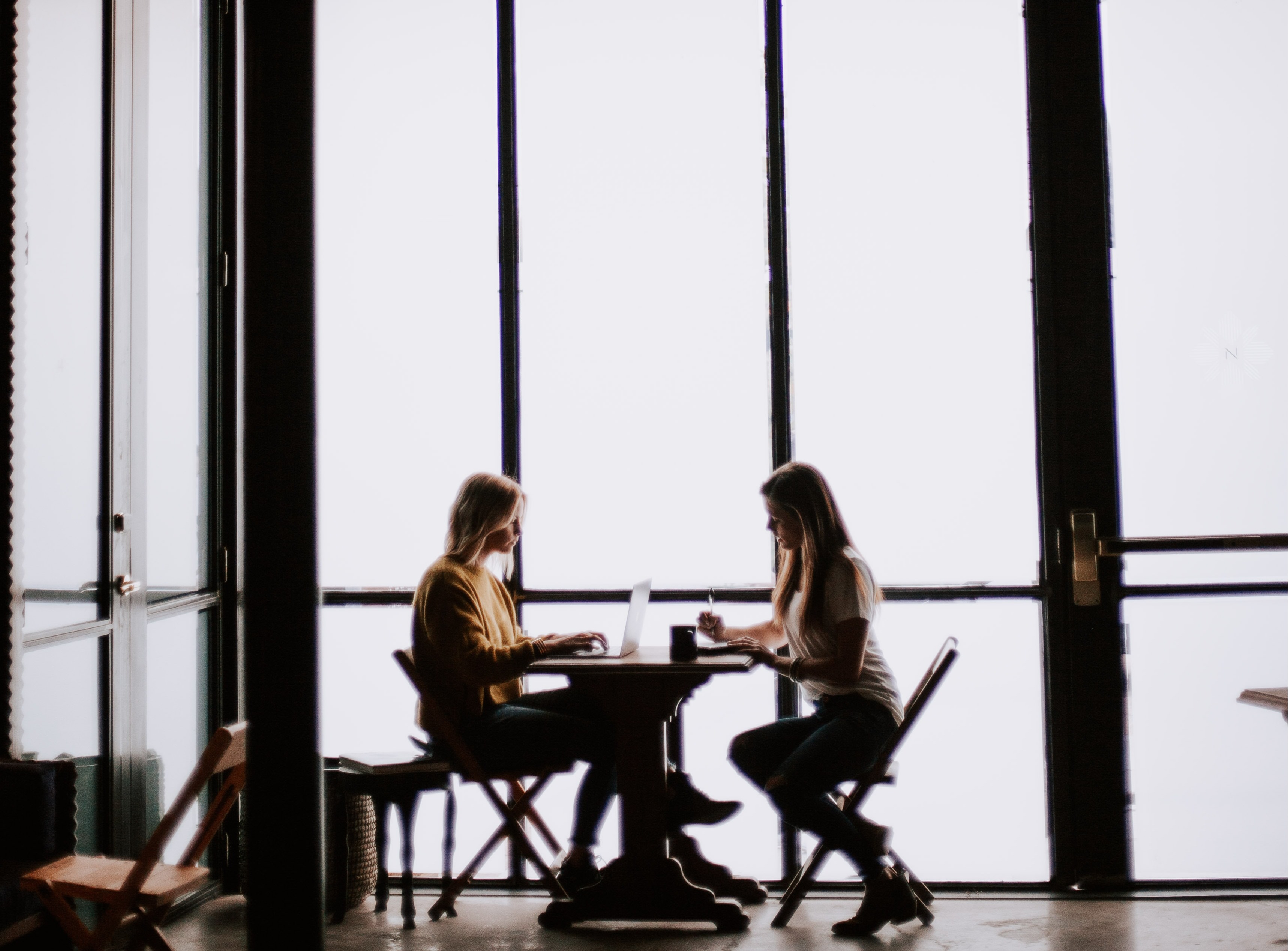 Two women sitting at a desk, one looking at her laptop screen and the other jotting down notes