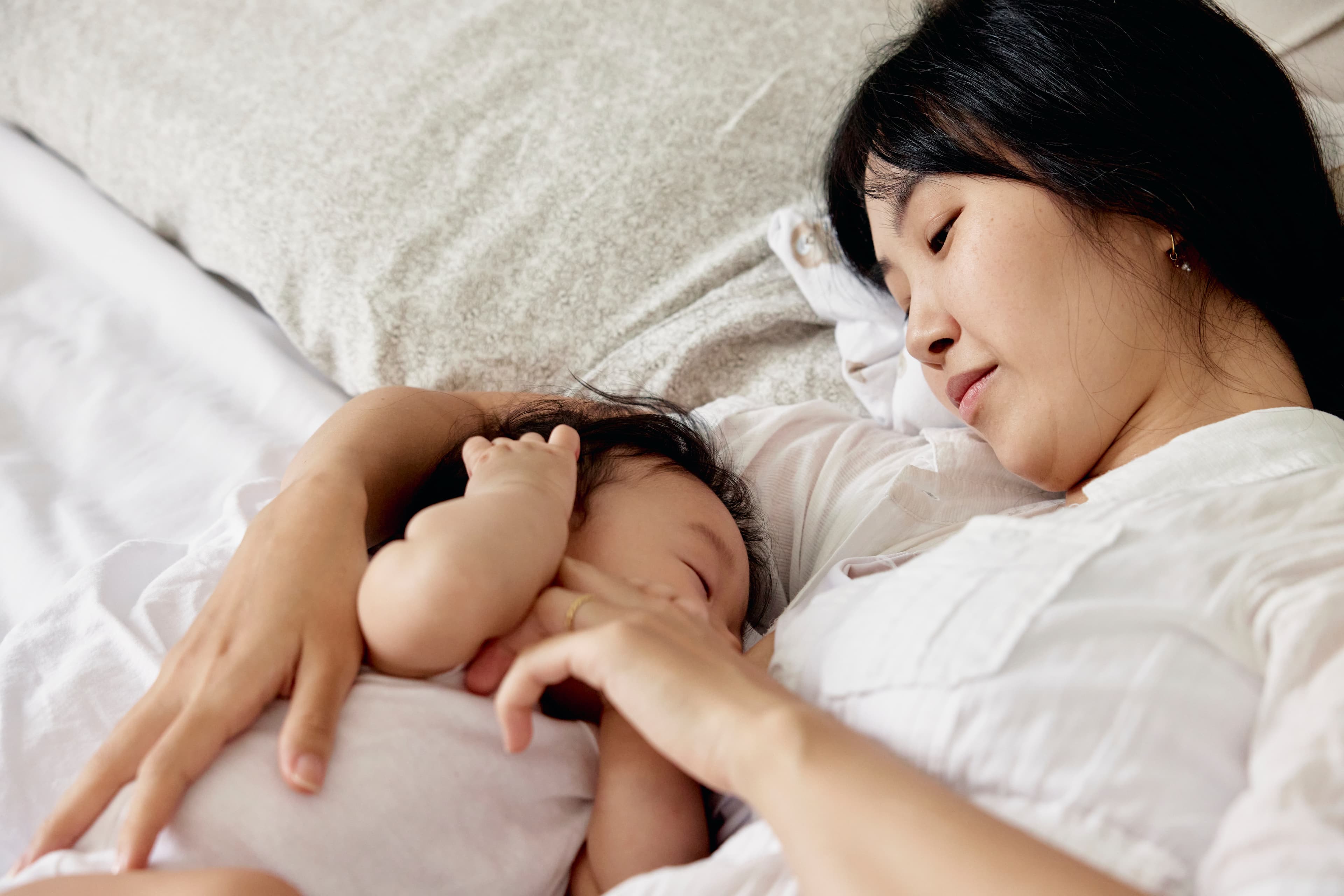 mother and baby lay in bed. mother is watching baby sleep and had her hand on the baby's cheek.
