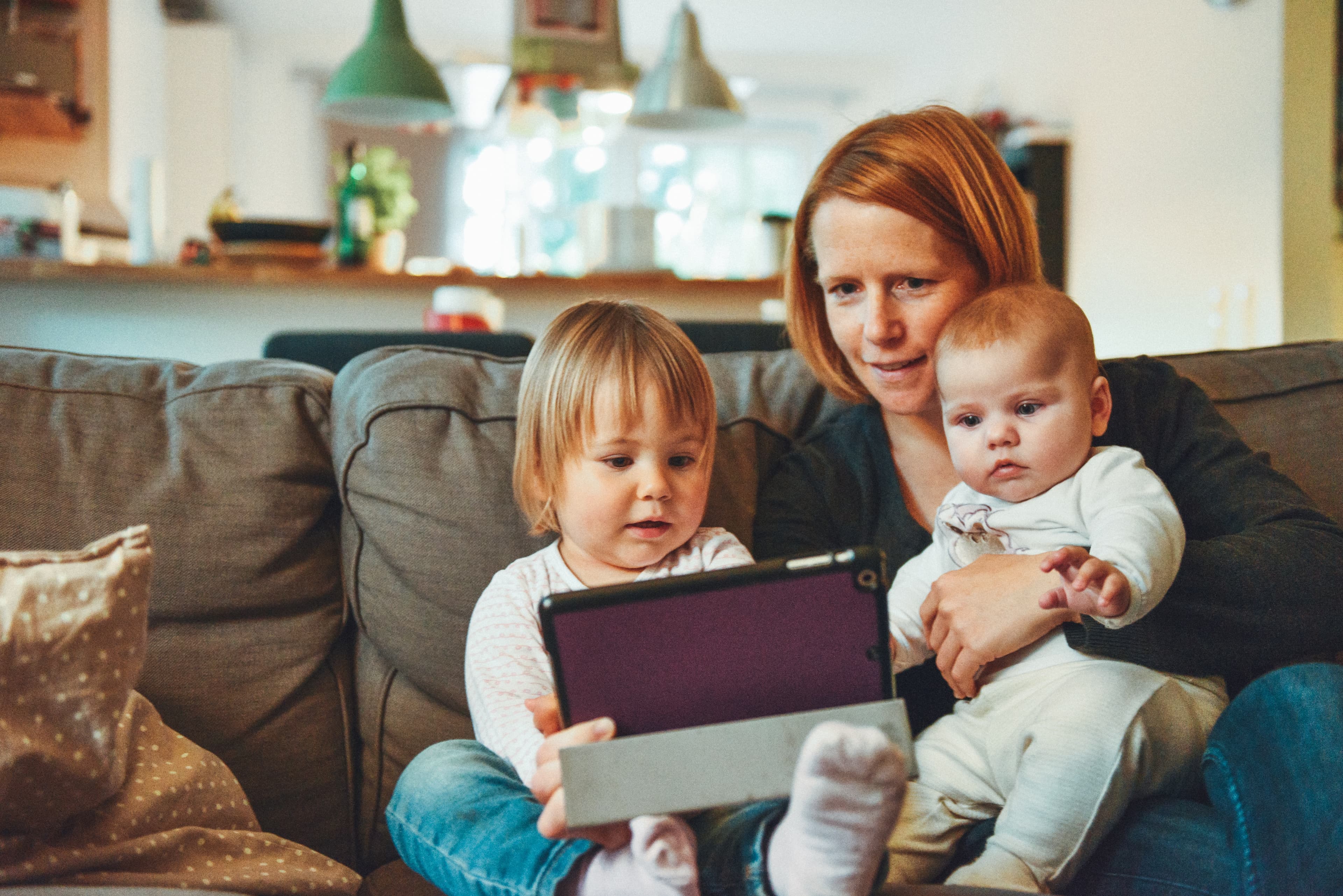 a mother holding her baby and toddler and sitting on a sofa in a living room. the mother is holding an ipad and looking at it with the children